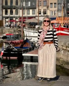 Elske, the blog owner, smiling on the harbour in Honfleur, France, with colourful fishing boats and historic waterfront buildings behind her in soft afternoon light.
